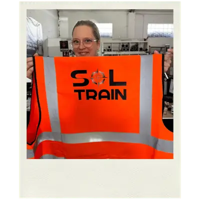 Polaroid-Foto einer Mitarbeiterin, die eine orangefarbene SOL TRAIN Warnweste hochhält